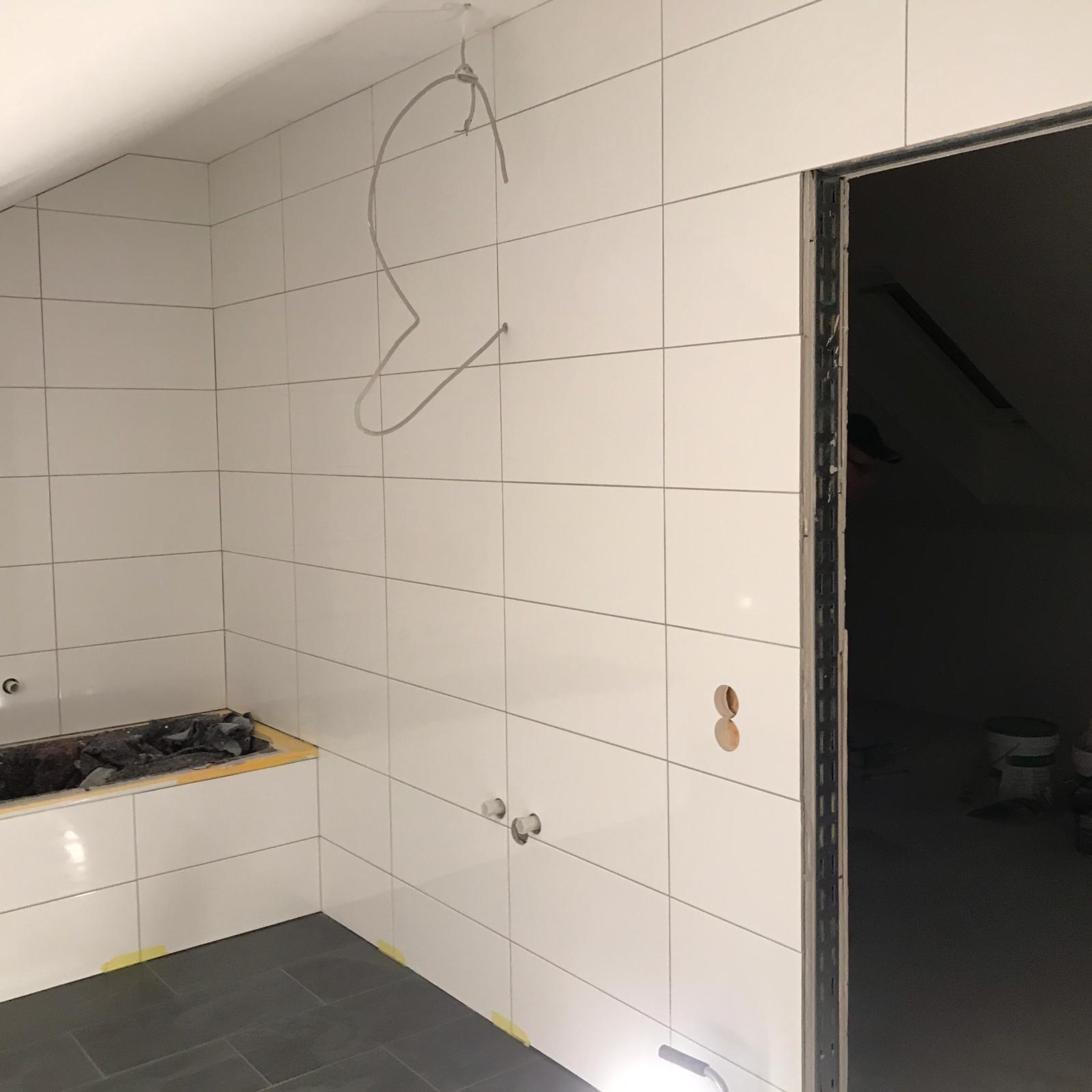 Wnętrze łazienki w trakcie prac: białe, błyszczące płytki ścienne ułożone do sufitu, ciemnoszara podłoga, przygotowane otwory na armaturę, widoczne przewody elektryczne i zabezpieczona wanna.