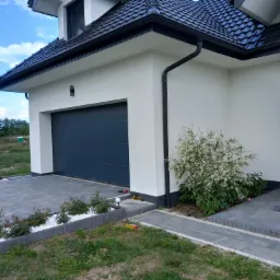 Nowoczesny dom jednorodzinny z antracytową bramą garażową, wybrukowanym podjazdem i schodami wejściowymi, otoczony zielenią z elementami dekoracyjnymi z białego kamienia.