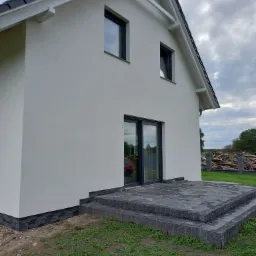 Nowoczesny dom z białymi ścianami i czarnymi oknami, widoczne dwustopniowe schody z szarych płyt prowadzące do drzwi tarasowych, w tle stos drewna.