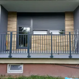 Zabudowany balkon z szarymi roletami zewnętrznymi, drewnopodobną okładziną ściany, metalową balustradą i widokiem na zieleń za oknem.