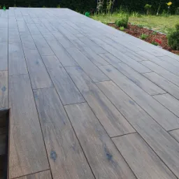 Nowoczesny taras wyłożony podłużnymi płytkami imitującymi drewno w odcieniach szarości i brązu, z widocznym fragmentem obrzeża i zielonym ogrodem w tle.