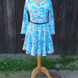 Dziecięca sukienka z długim rękawem w niebiesko-biały kwiatowy wzór, przewiązana czarnym paskiem, prezentowana na drewnianym manekinie na tle ciemnego, drewnianego płotu.