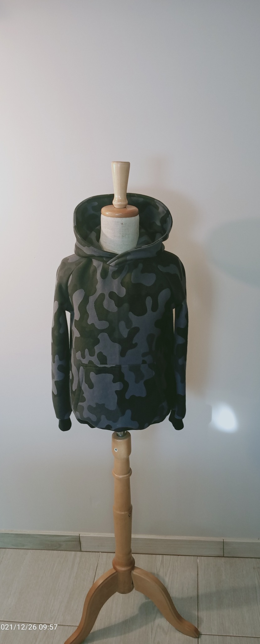 Dziecięca bluza z kapturem w kamuflażu, prezentowana na drewnianym manekinie, na tle białej ściany i jasnej podłogi.