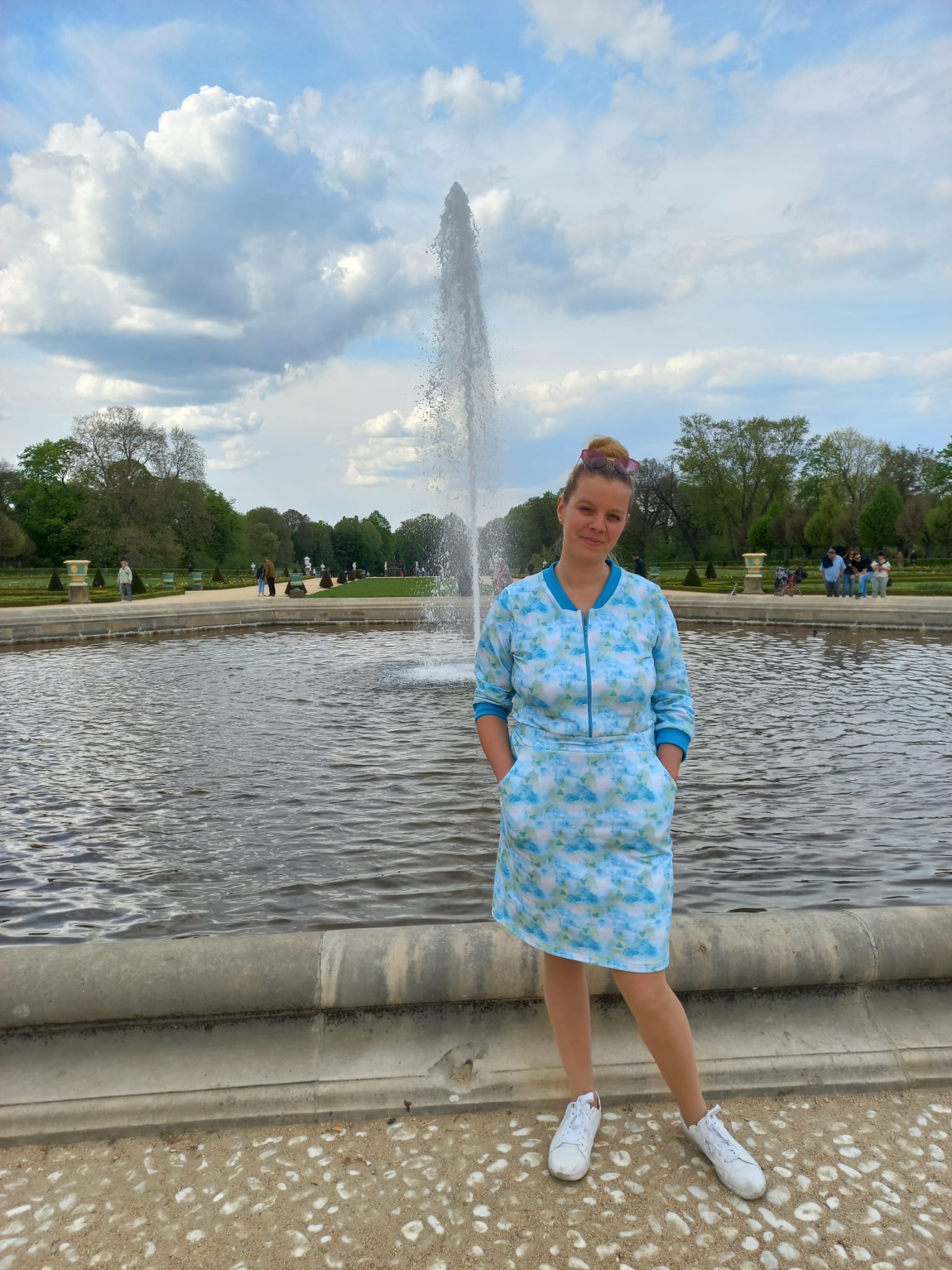 Kobieta w samodzielnie uszytej, niebieskiej sukience w białe wzory, stojąca na tle fontanny w parku.