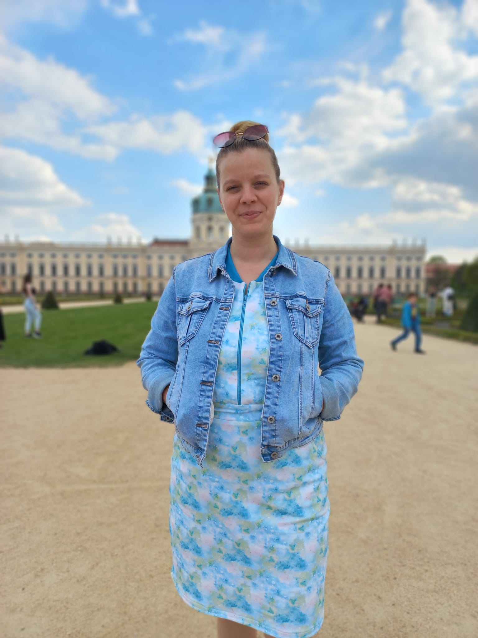 Kobieta w dżinsowej kurtce i wzorzystej sukience na tle pałacowego ogrodu.