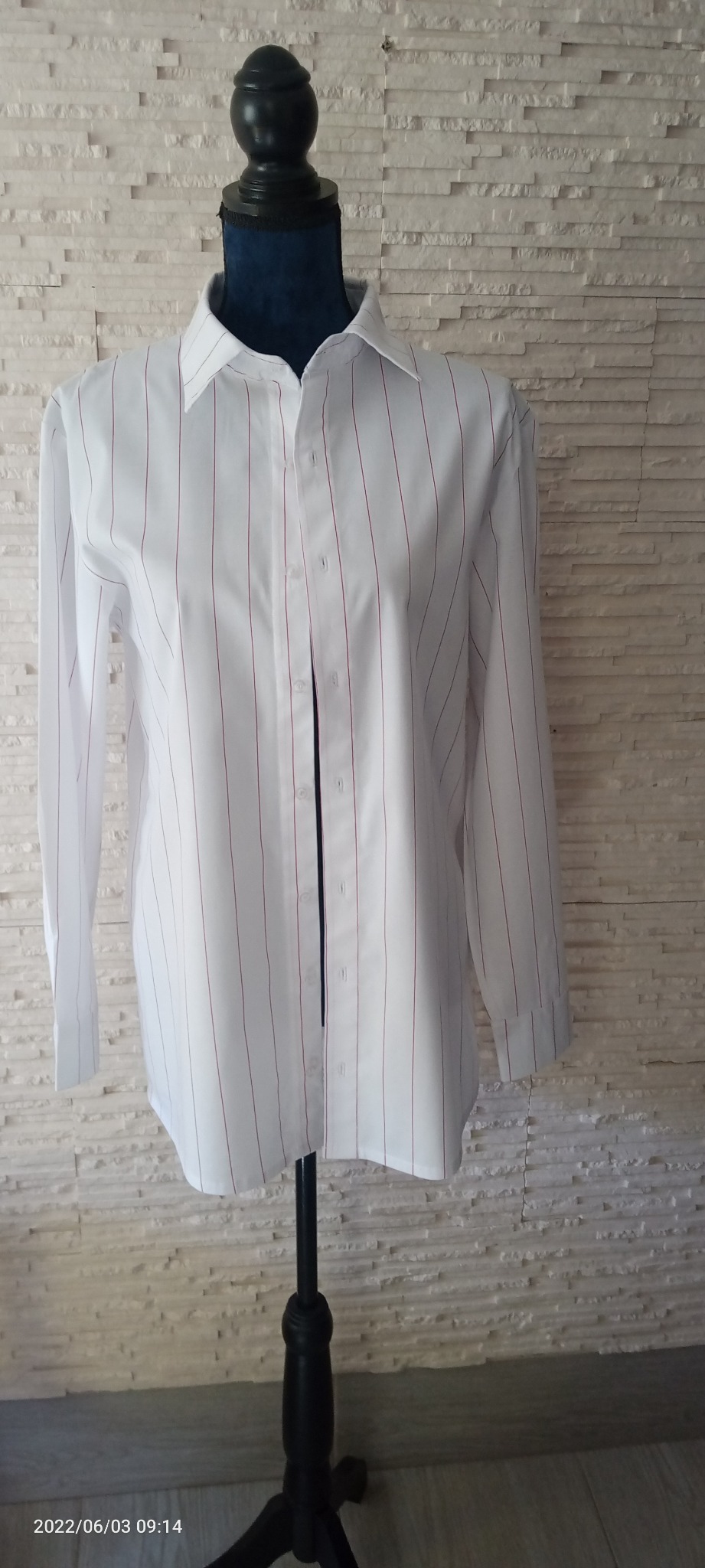 Biała koszula w cienkie czerwone paski, rozpięta, prezentowana na czarnym manekinie na tle ściany z białych paneli imitujących kamień.