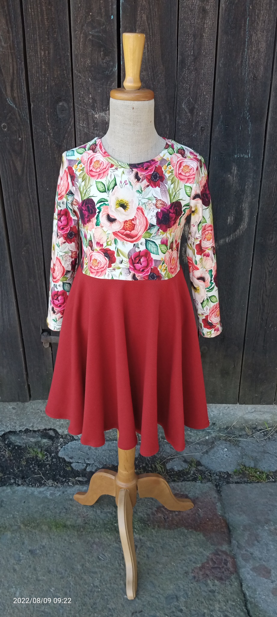 Dziewczęca sukienka z długim rękawem, z motywem kwiatowym na górze i gładką, czerwoną spódnicą, prezentowana na drewnianym manekinie na tle ciemnych, drewnianych desek.