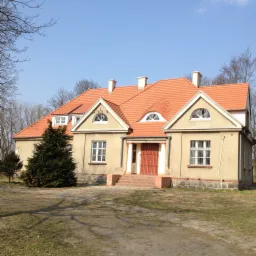 Jasny, parterowy dom z pomarańczową dachówką, widoczne dwa kominy i półokrągłe okna na poddaszu, na pierwszym planie iglak