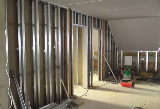 Wnętrze w trakcie prac: widoczne metalowe profile ścian działowych, częściowo zabudowany sufit poddasza, nagrzewnica i przewody elektryczne na podłodze.