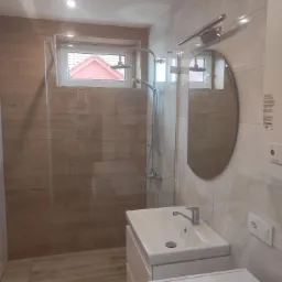 Nowoczesna łazienka z beżowymi płytkami imitującymi drewno, kabiną prysznicową typu walk-in, owalnym lustrem, białą umywalką z szafką i pralką pod blatem.