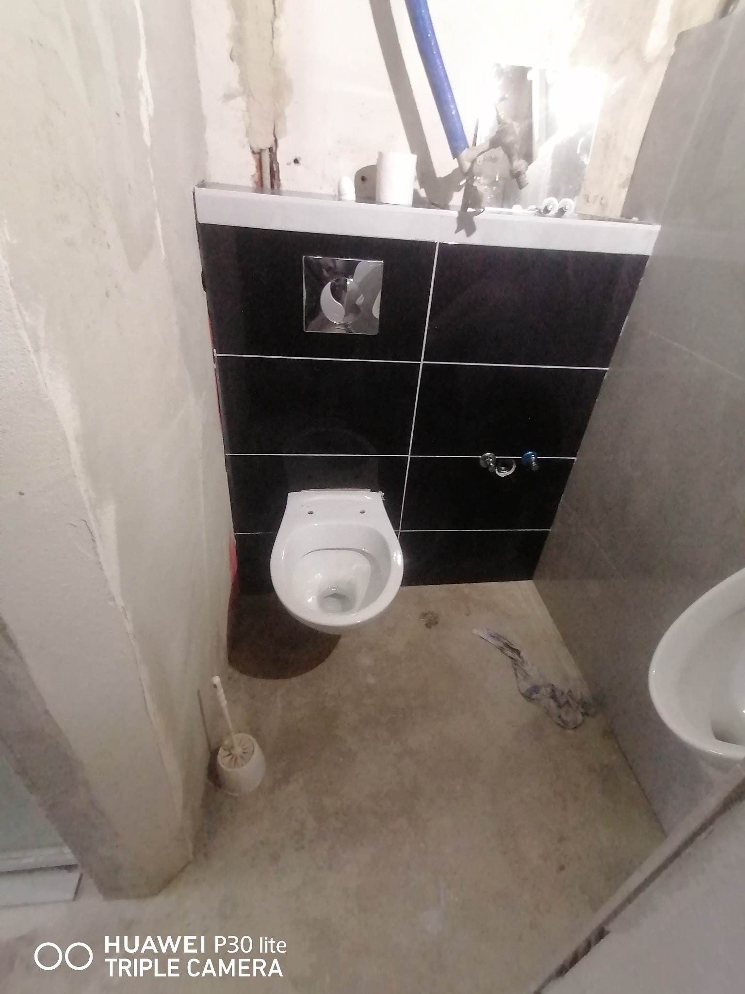 Wnętrze łazienki w trakcie prac: widoczna podwieszana toaleta z czarną, kwadratową płytką za nią, surowe ściany przygotowane do dalszych prac wykończeniowych, oraz szczotka do WC. Na podłodze...