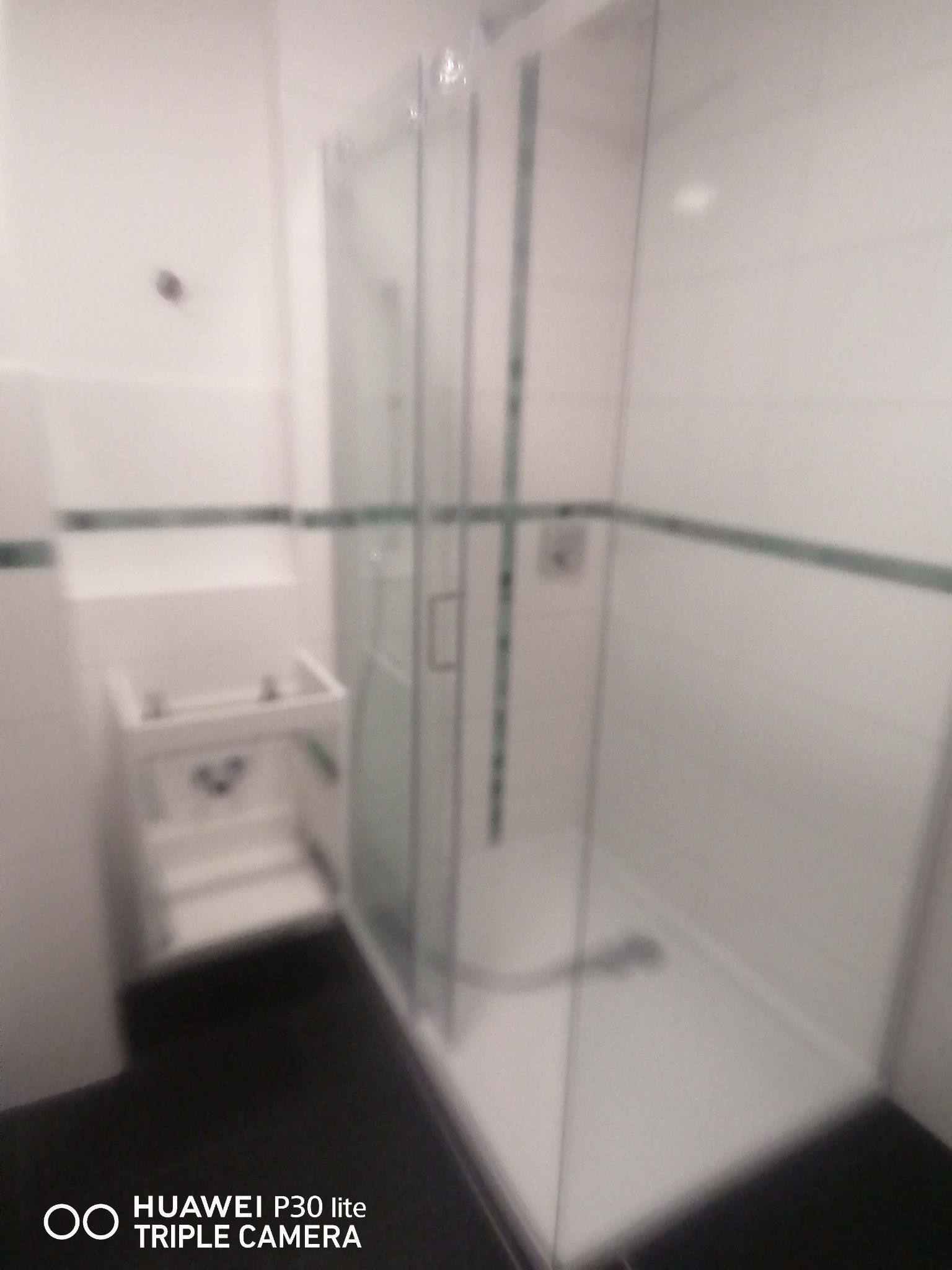 Biała łazienka z kabiną prysznicową ze szklanymi drzwiami, umywalką i czarną podłogą, wykończona płytkami z zielonym paskiem dekoracyjnym.