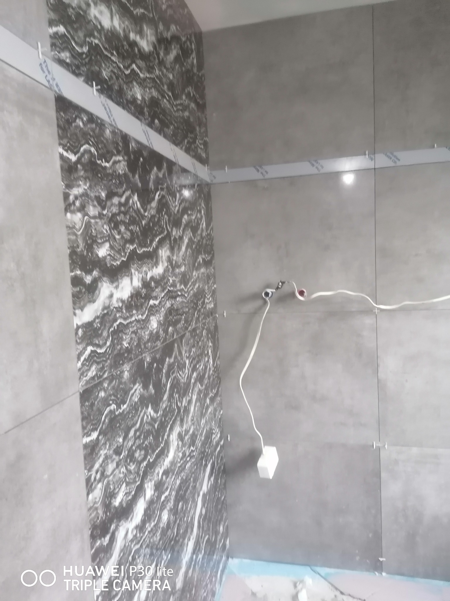 Ściana łazienki w trakcie układania płytek imitujących marmur, z widocznymi listwami poziomującymi, odstającymi kablami elektrycznymi i krzyżykami dystansowymi.