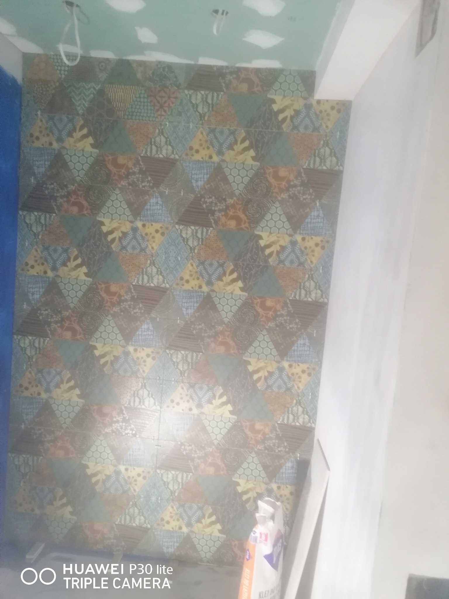 Ściana wyłożona dekoracyjnymi, trójkątnymi płytkami w różnych wzorach i kolorach, z widocznym workiem kleju do płytek w rogu pomieszczenia.