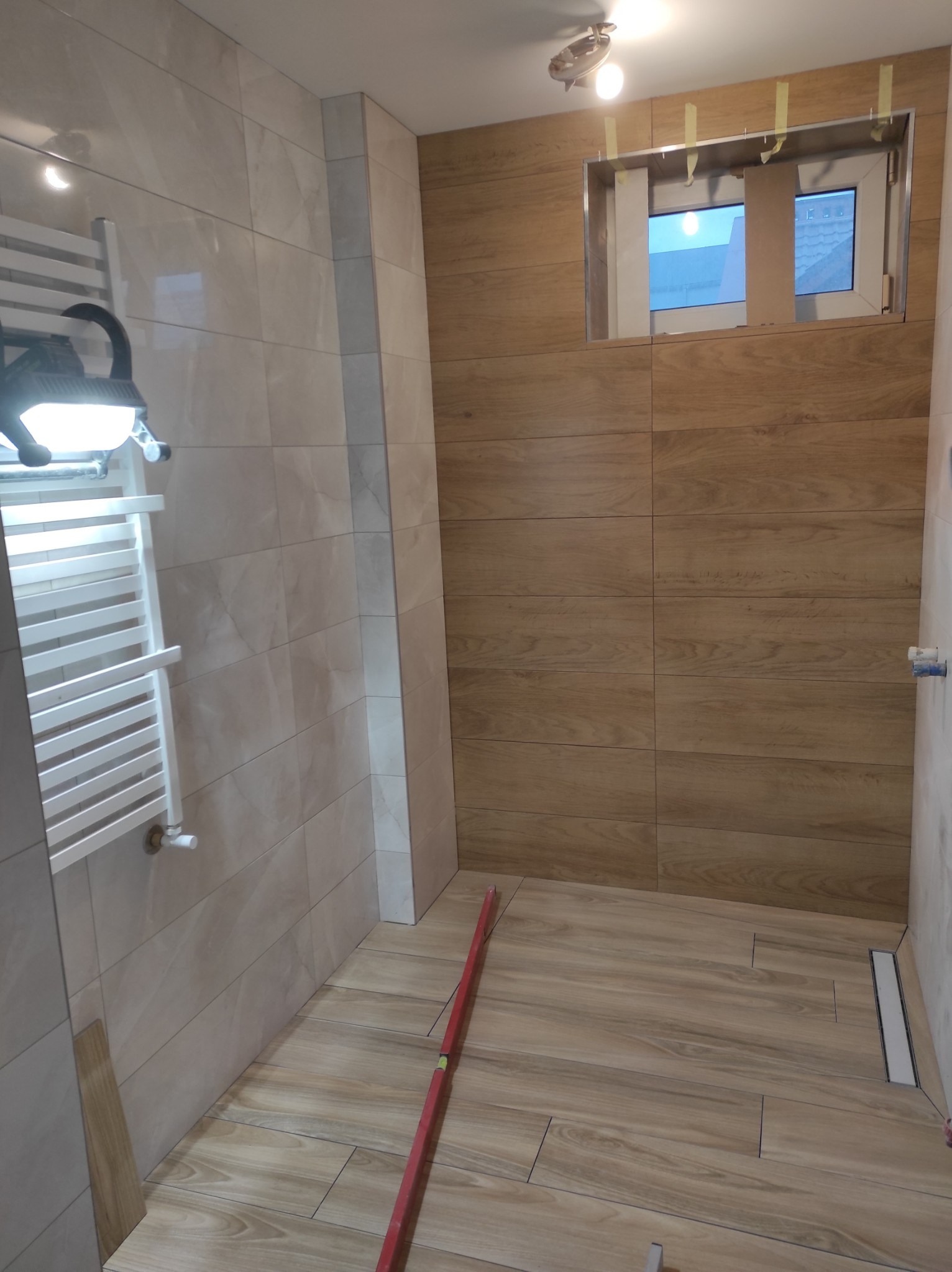 Ułożone płytki imitujące drewno na ścianach i podłodze w łazience, widoczny grzejnik drabinkowy, poziomica na podłodze i okno z taśmą malarską.