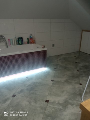 Wnętrze łazienki na poddaszu z wanną obudowaną mozaiką z podświetleniem LED, szare płytki podłogowe z ciemnymi wstawkami, białe płytki ścienne, skośny sufit.