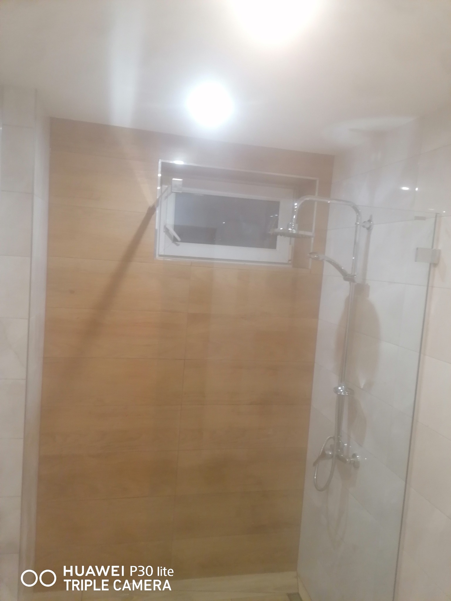 Nowoczesna kabina prysznicowa z deszczownicą i przeszklonymi drzwiami, obok ściana wyłożona płytkami imitującymi drewno z małym oknem.