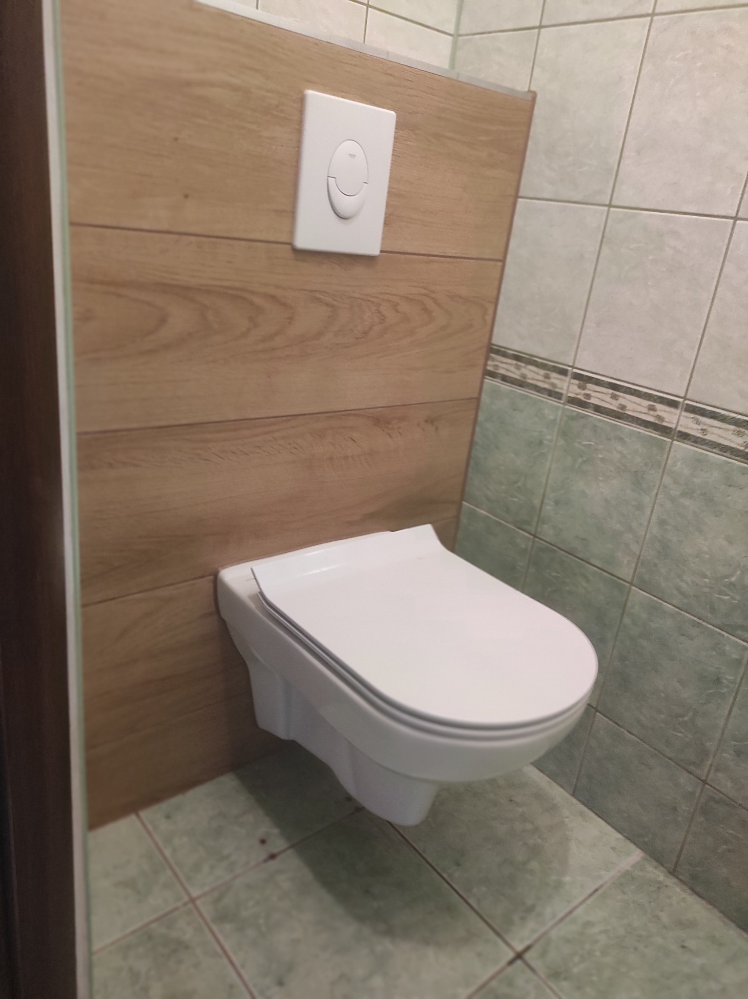 Wisząca miska WC z białą deską i przyciskiem spłukującym na ścianie wyłożonej panelami imitującymi drewno, obok ściana z zielonych płytek.