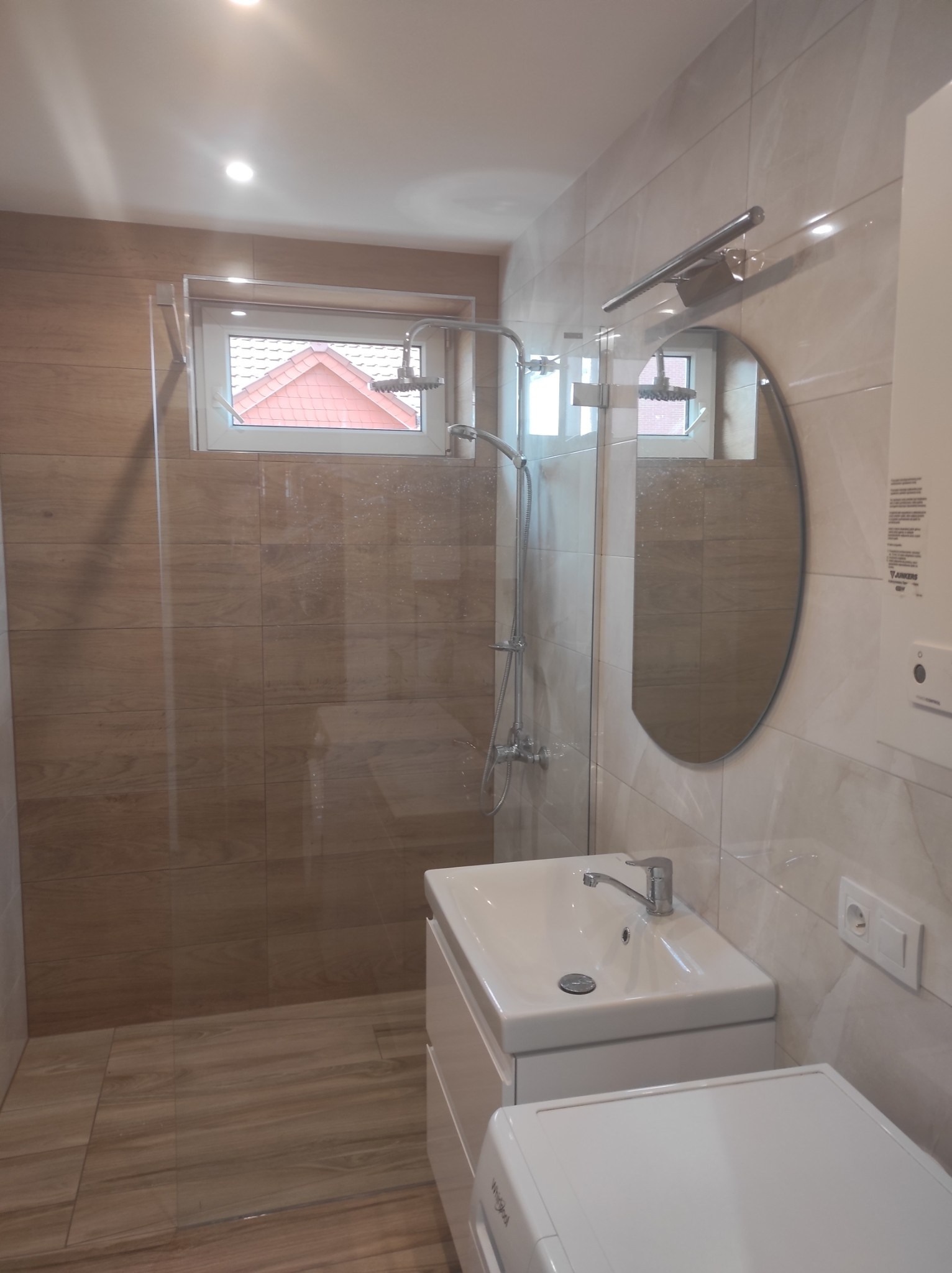 Nowoczesna łazienka z beżowymi płytkami imitującymi drewno, kabiną prysznicową typu walk-in, owalnym lustrem, białą umywalką z szafką i pralką pod blatem.