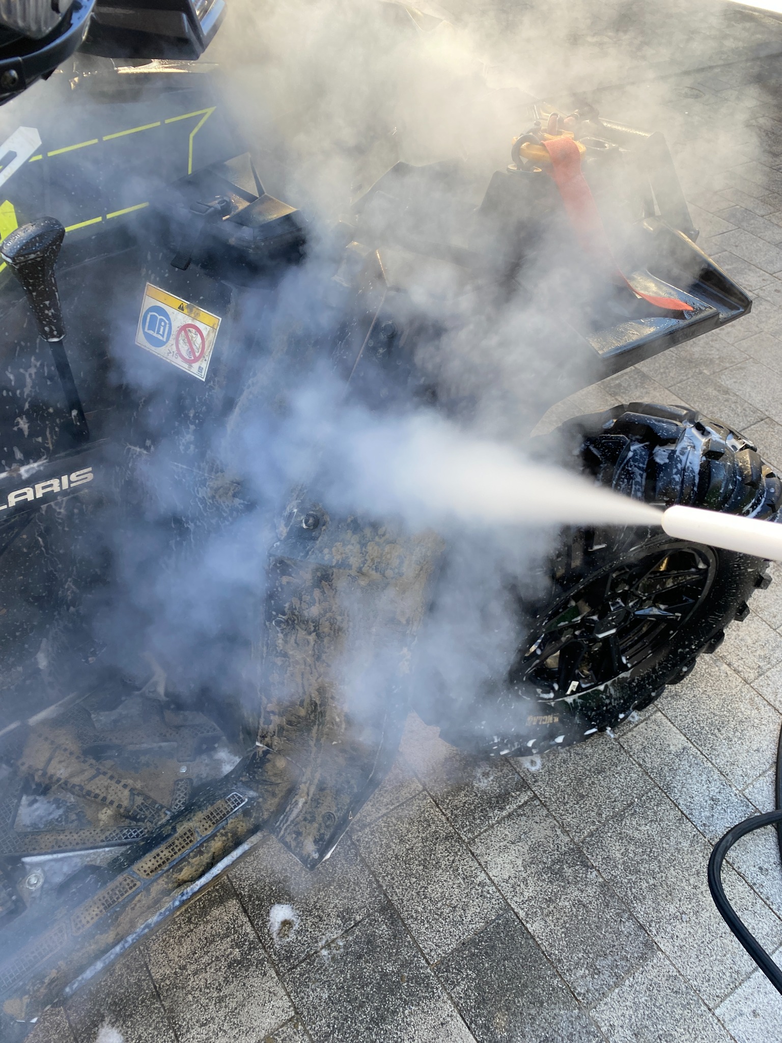 Czyszczenie quada marki Polaris z błota za pomocą myjki ciśnieniowej, widoczne silne strumienie wody i para.