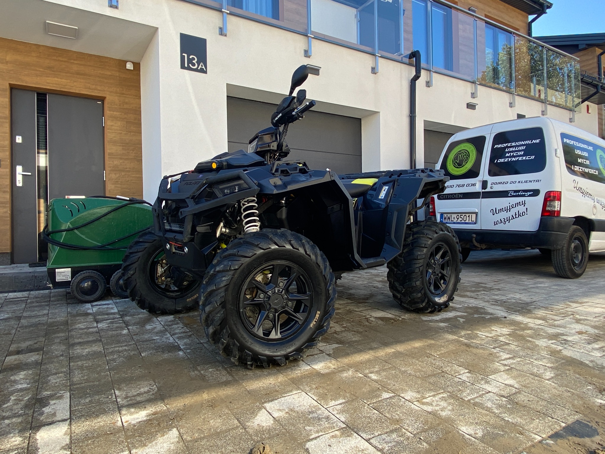 Czarny quad Polaris stoi przed budynkiem z numerem 13A, obok zaparkowany biały Citroen Berlingo z reklamą usług mycia i dezynfekcji.