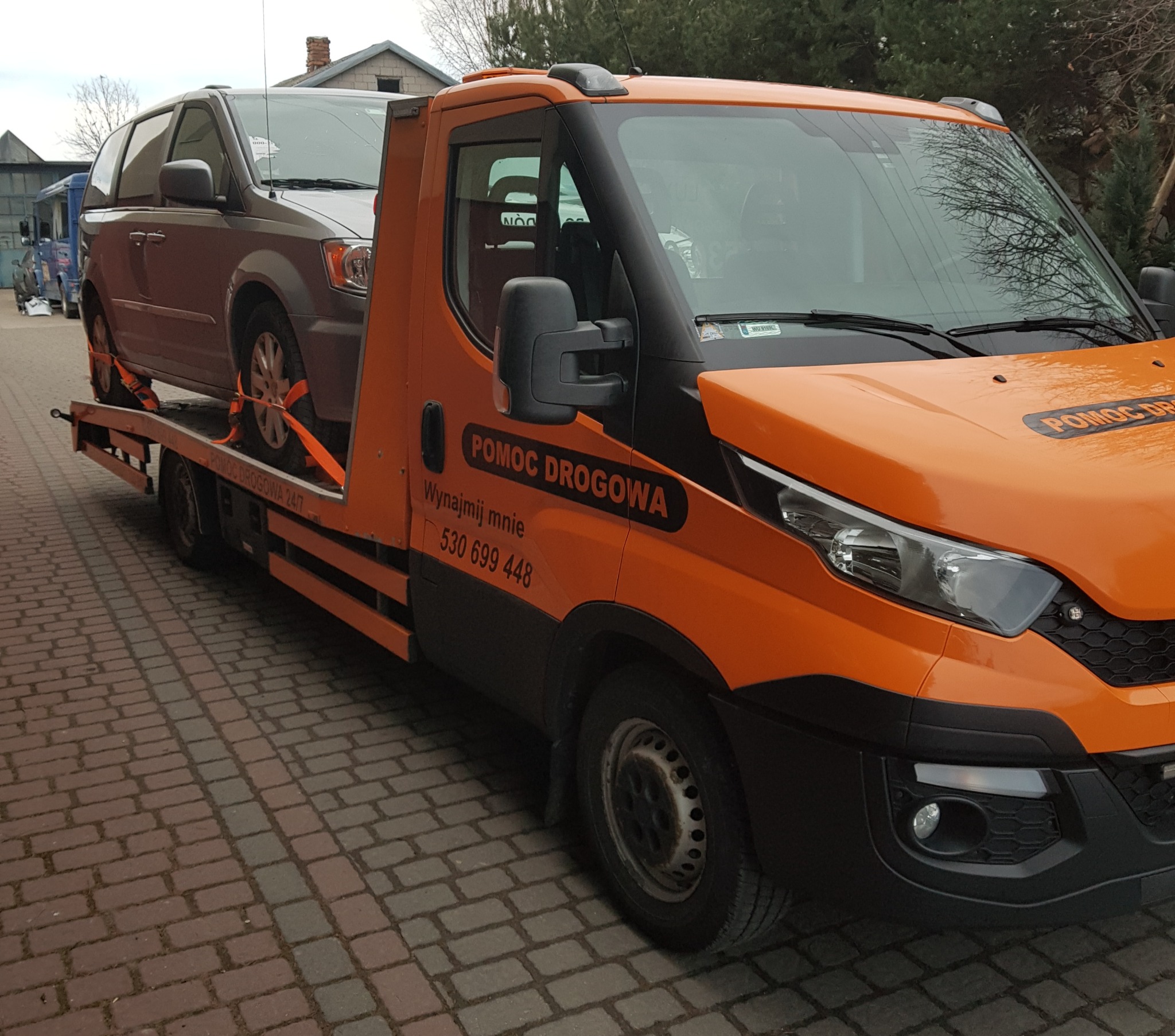 Pomarańczowa laweta Iveco z szarym minivanem zabezpieczonym pomarańczowymi pasami transportowymi, na brukowanym podjeździe. Na kabinie lawety napis 'POMOC DROGOWA' i numer telefonu.