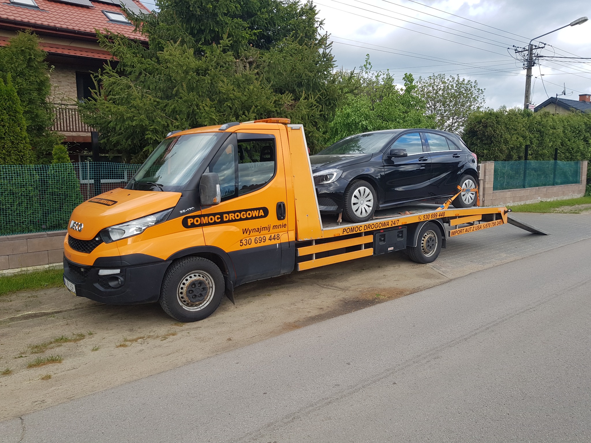 Pomarańczowa laweta Iveco z czarnym Mercedesem klasy A na platformie, zabezpieczonym pasami transportowymi, na tle zieleni i domu jednorodzinnego.
