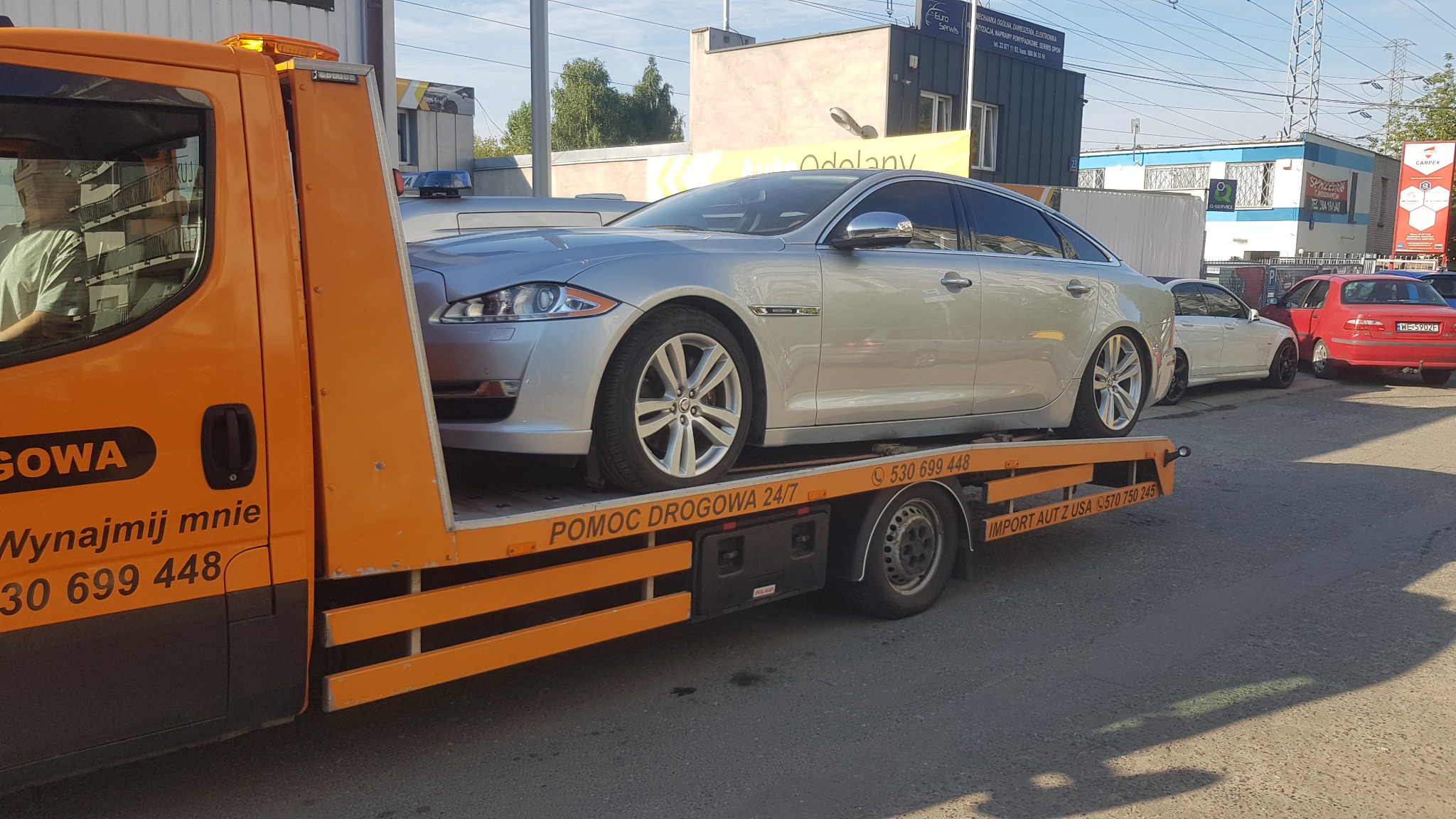 Pomarańczowa laweta z srebrnym Jaguarem XJ na platformie, widoczny numer telefonu kontaktowego i napis POMOC DROGOWA 24/7, w tle zabudowa miejska.