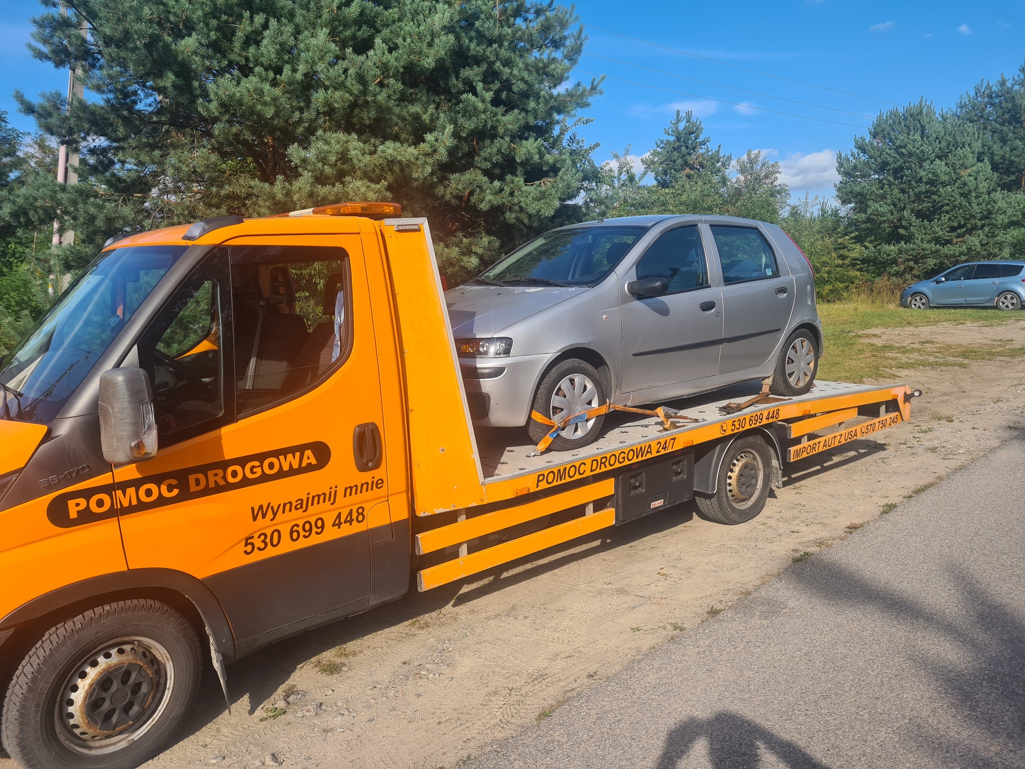 Żółta laweta z srebrnym samochodem osobowym na platformie, zabezpieczonym pomarańczowymi pasami, w tle drzewa iglaste i błękitne niebo.