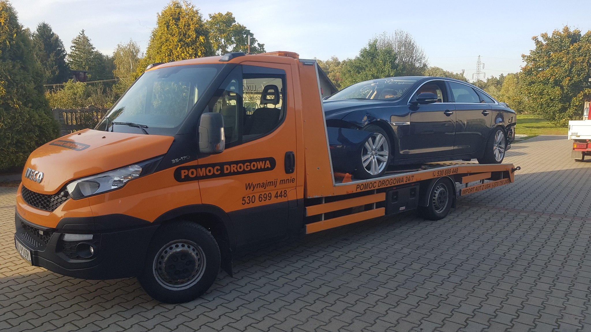 Pomarańczowa laweta Iveco z uszkodzonym, ciemnym sedanem marki Jaguar na platformie, zaparkowana na brukowanym placu, widoczne numery kontaktowe i logo firmy Pomoc Drogowa.
