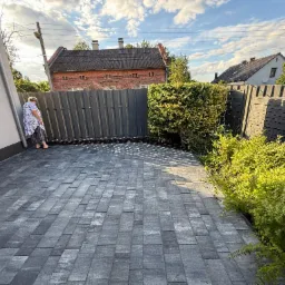 Ułożona szara kostka brukowa na podjeździe, widok z góry, z boku ściana budynku i ogrodzenie z zielenią. Kobieta oparta o ścianę, w tle budynki.