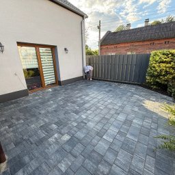 BRUKUJEMY.PL SPÓŁKA Z OGRANICZONĄ ODPOWIEDZIALNOŚCIĄ - Szara kostka brukowa ułożona na podwórku przy domu z szarą elewacją. W tle widoczny płot i budynek z czerwonej cegły. Osoba pochylona nad krawędzią kostki.