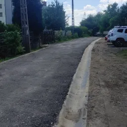 Nowo wybudowana droga asfaltowa z betonowym korytkiem odwadniającym po prawej stronie, widoczne słupy energetyczne i drzewa w tle, samochody zaparkowane w oddali.