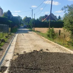 Przygotowanie podłoża pod drogę: wyrównany teren z nasypem ziemi, widoczny betonowy rów odwadniający, w tle domy i drzewa w słoneczny dzień.