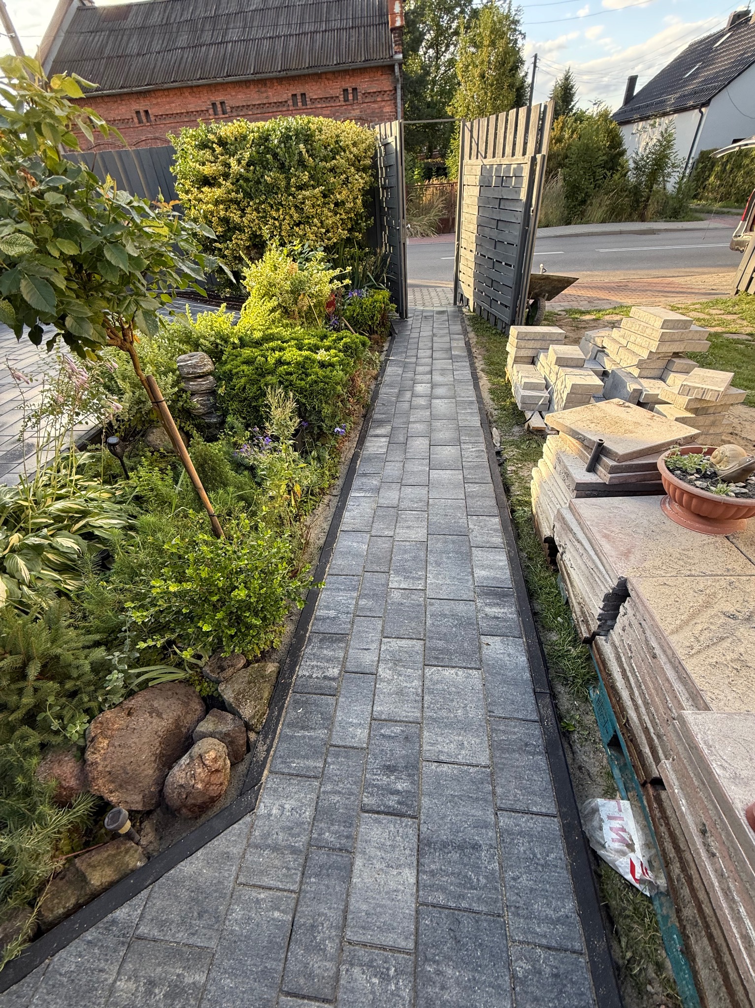 Ścieżka z kostki brukowej w Zabrzu, prowadząca do bramy. Po bokach rabaty z roślinami, po prawej stos kostki brukowej. Widok w perspektywie.