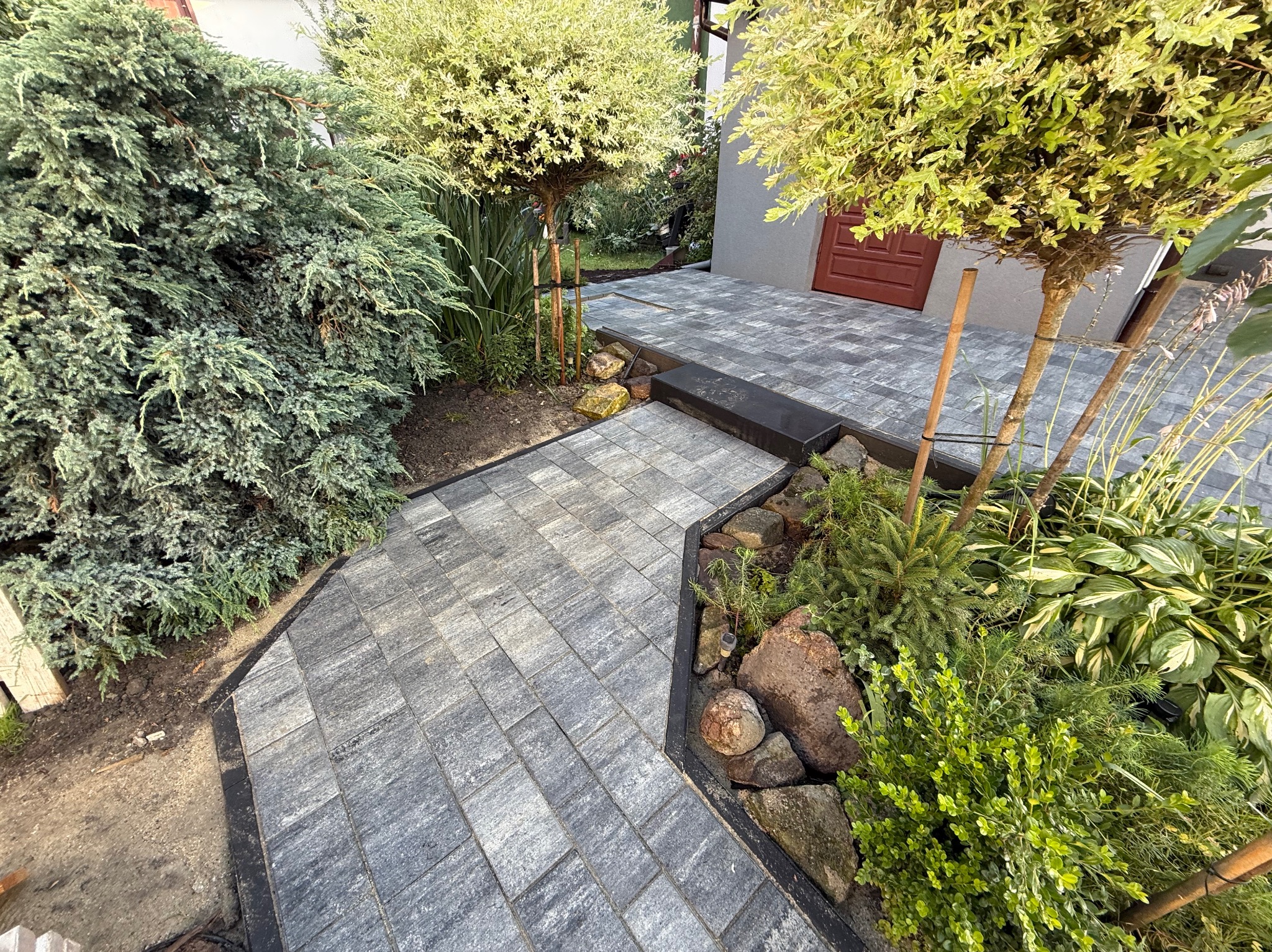 Ścieżka z kostki brukowej w ogrodzie z roślinnością, prowadząca do drzwi budynku. Obrzeża w kolorze czarnym, kostka szara. Widok z góry.