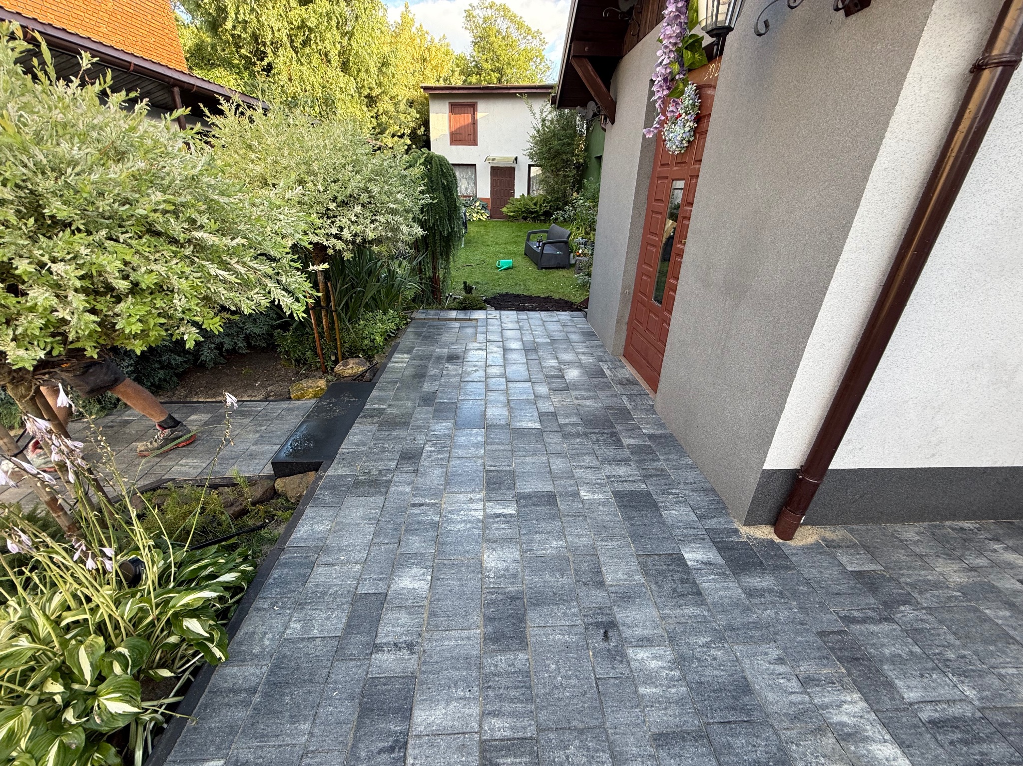 Ułożona szara kostka brukowa prowadząca od domu do ogrodu, z widoczną zielenią i częściowo widoczną osobą w lewym dolnym rogu kadru. Kostka ułożona równo, wzór prostokątny.