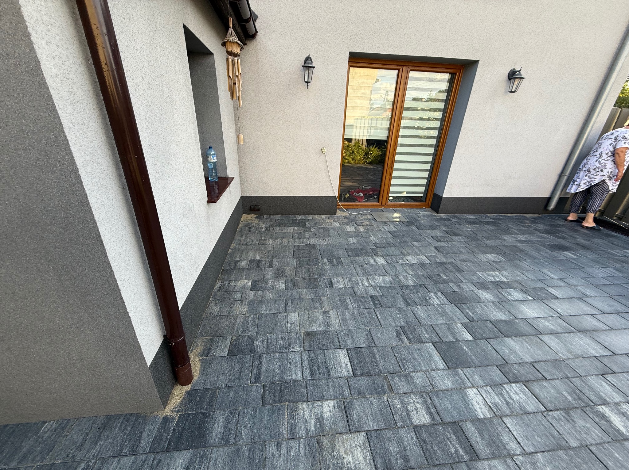 Ułożona, szara kostka brukowa przed budynkiem z brązową rynną i elementami dekoracyjnymi. Widoczna butelka wody na parapecie. Fragment osoby w prawym rogu kadru.