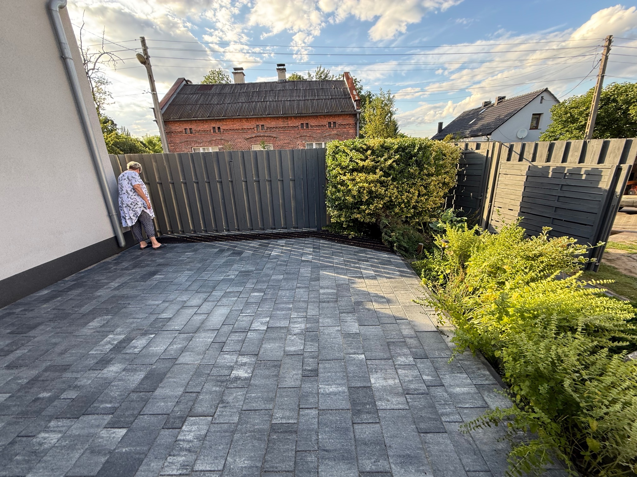 Ułożona szara kostka brukowa na podjeździe, widok z góry, z boku ściana budynku i ogrodzenie z zielenią. Kobieta oparta o ścianę, w tle budynki.