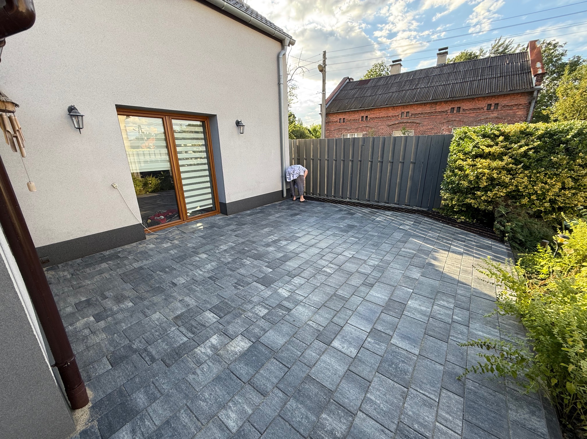 Szara kostka brukowa ułożona na podwórku przy domu z szarą elewacją. W tle widoczny płot i budynek z czerwonej cegły. Osoba pochylona nad krawędzią kostki.