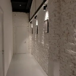 Korytarz z białą, nier&oacute;wną ścianą z cegły, oświetloną punktowymi lampami sufitowymi, z trzema obrazami w czarnych ramkach wiszącymi na ścianie, gładką białą podłogą i białymi drzwiami po obu stronach.