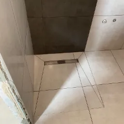 Ułożone płytki podłogowe i ścienne w łazience, widoczny odpływ liniowy w strefie prysznica, fragment surowego wykończenia przy framudze drzwi.