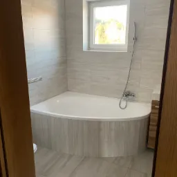 Jasna łazienka po remoncie z wanną w zabudowie i oknem z widokiem na zieleń, płytki imitujące marmur na ścianach i podłodze, widok zza framugi drzwi.