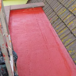 Czerwona, świeżo nałożona posadzka żywiczna na dachu płaskim, widok z g&oacute;ry z drabiny, obok dach&oacute;wka z mchem i fragment okna dachowego.