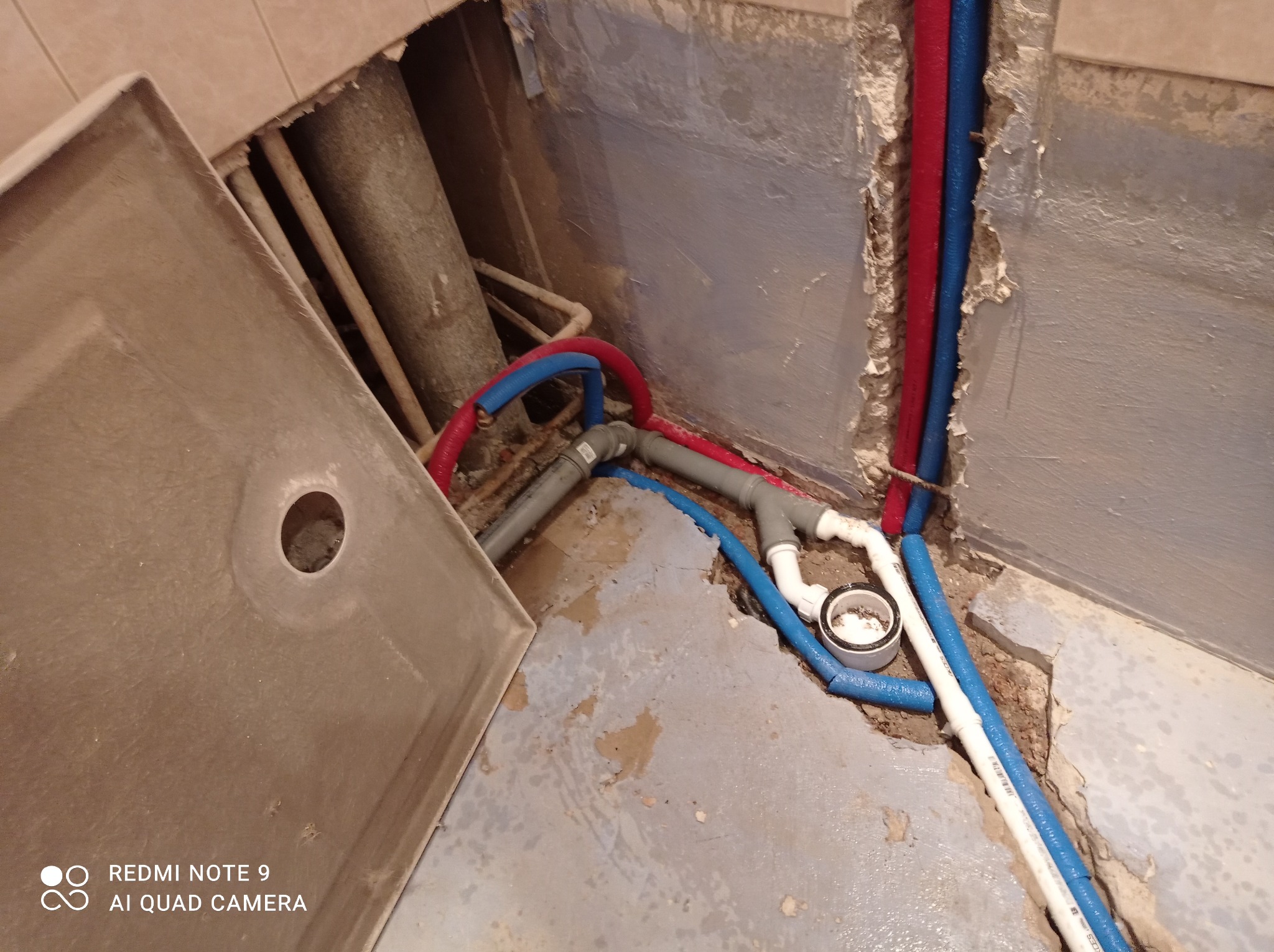Niedokończona instalacja wodno-kanalizacyjna w łazience, widoczne rury czerwone i niebieskie prowadzone w bruzdach w ścianie, szara podłoga z resztkami zaprawy, białe i szare rury PCV z kształtkami...