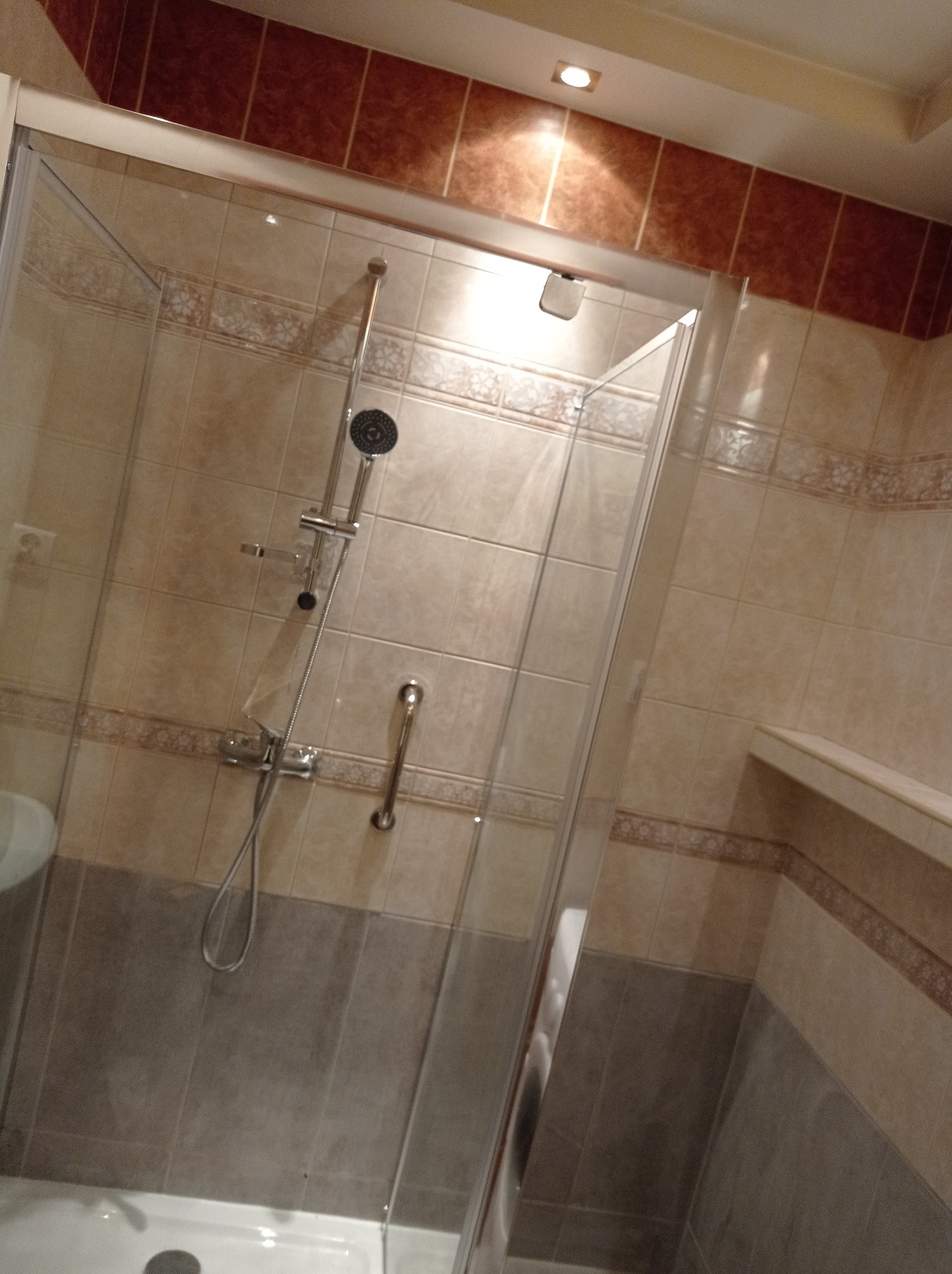 Zamknięta kabina prysznicowa z chromowaną armaturą, beżowe i brązowe płytki z dekoracyjnym pasem, widoczny fragment umywalki i wnęka w ścianie.