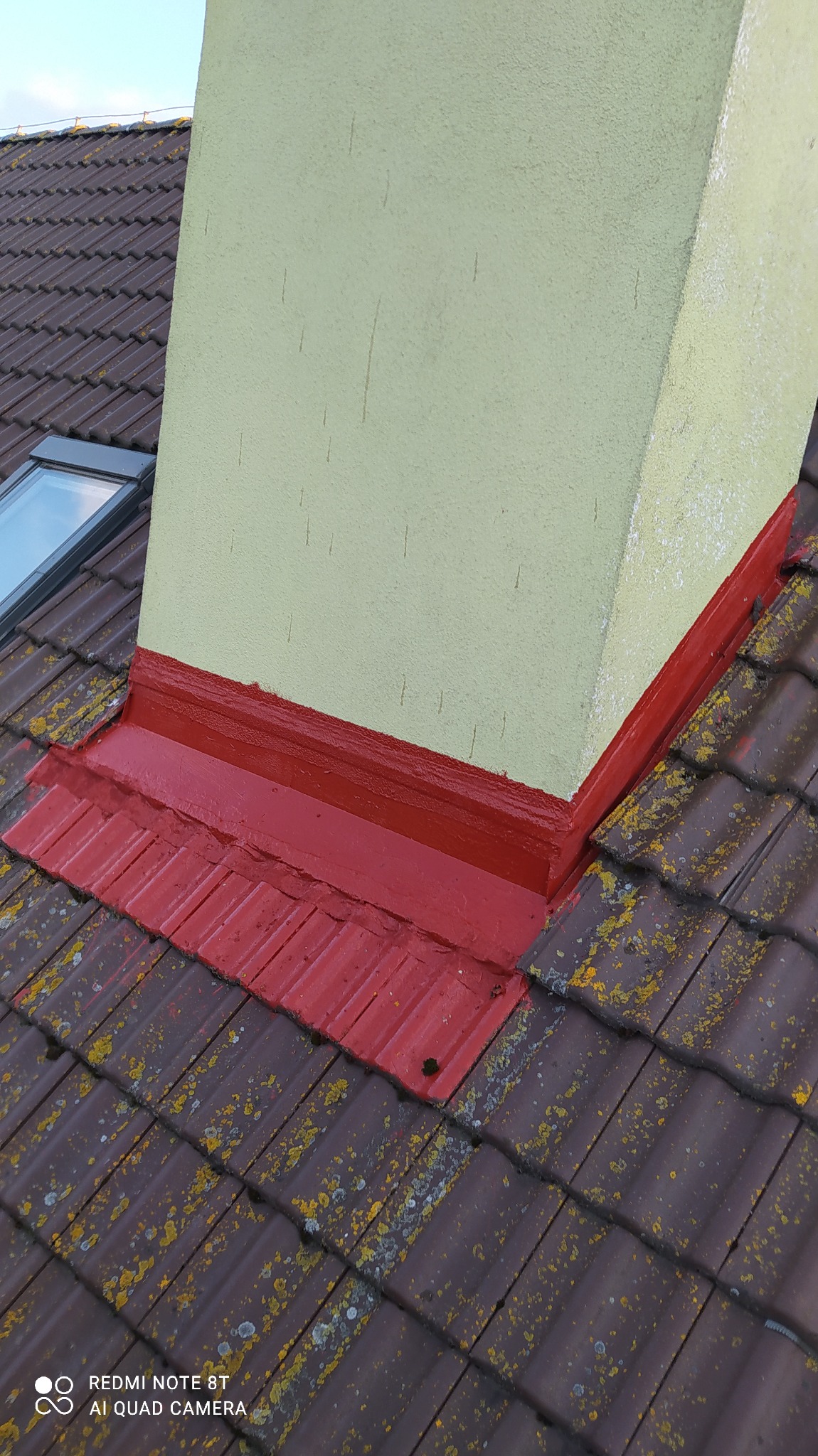 Otynkowany komin pomalowany na jasnożółty kolor, z czerwonym obramowaniem u podstawy, wystający z dachu pokrytego brązową dachówką z widocznymi porostami.