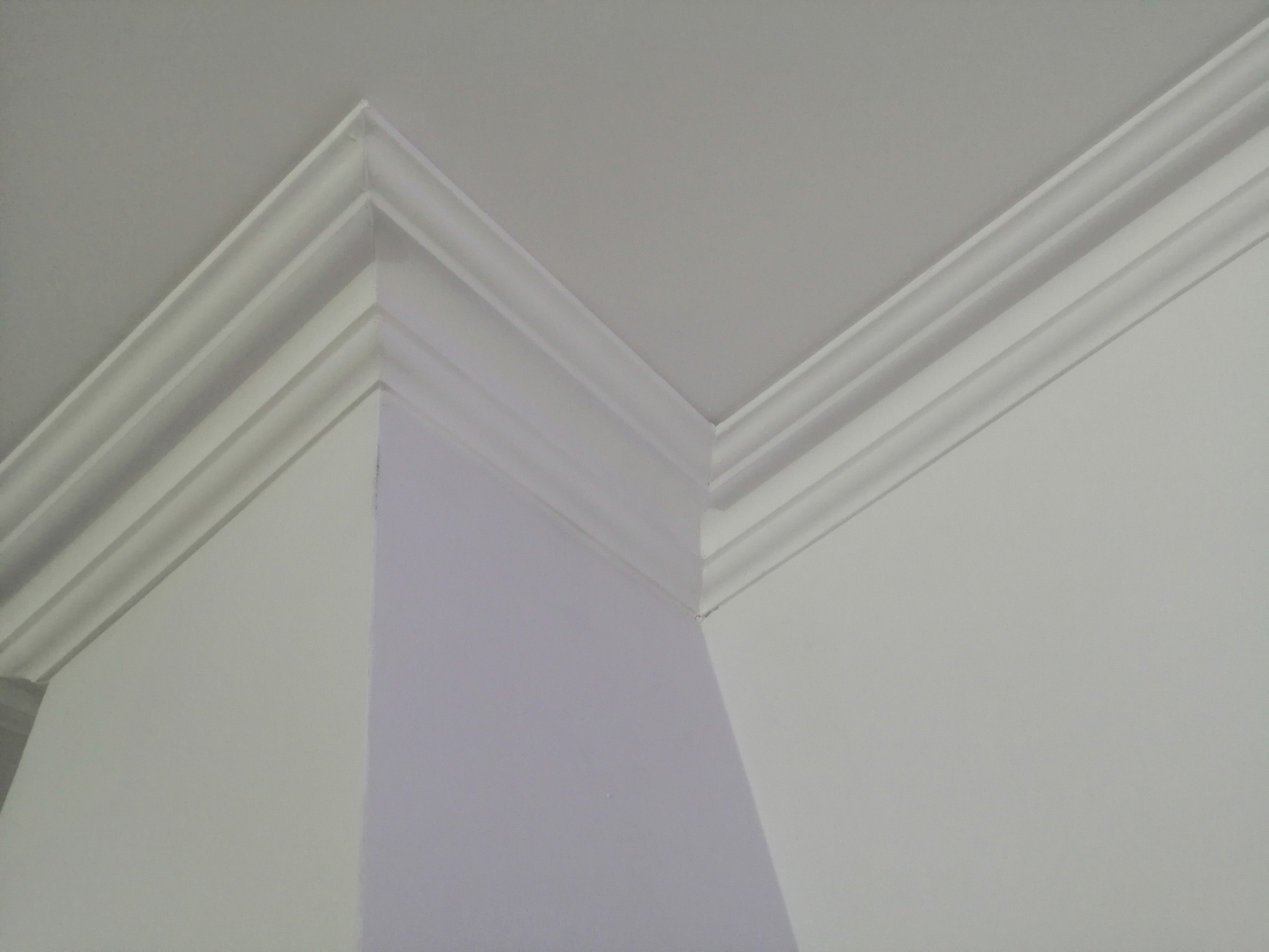 Wykończony narożnik sufitu z ozdobną, białą listwą przysufitową, widok z dołu na połączenie dwóch ścian w jasnym, neutralnym wnętrzu.