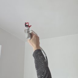 BIG-MARK - Malowanie natryskowe sufitu w pomieszczeniu, widoczny fragment twarzy malarza z zabezpieczeniem na głowie, pistolet natryskowy w dłoni oraz zaklejone miejsce po lampie.