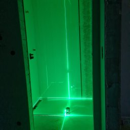 BIG-MARK - Wnętrze łazienki w trakcie remontu, oświetlone zielonym laserem krzyżowym, wyznaczającym poziomy na ścianach pokrytych płytkami i podłodze. Widoczny miernik laserowy i zwinięta miarka.